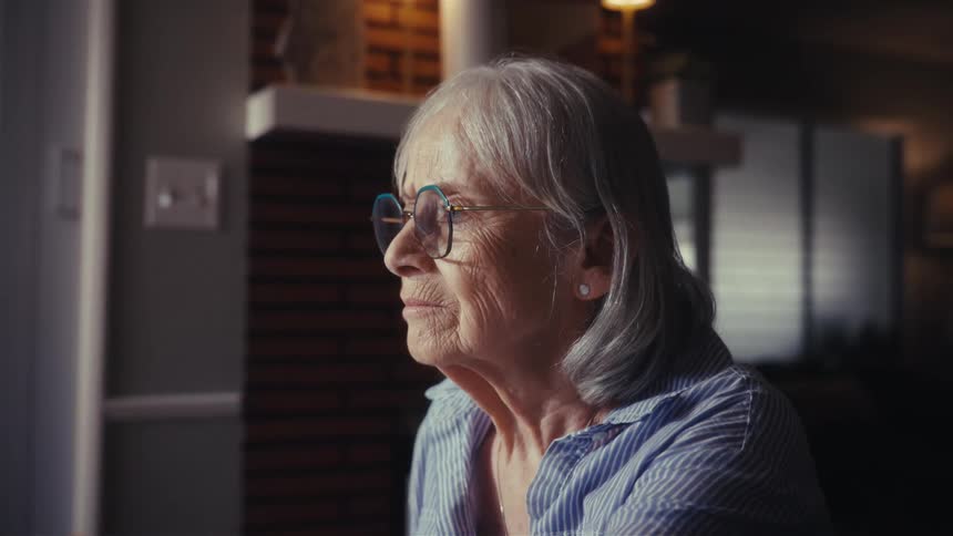 A concerned elderly woman with glasses sits at a table in a dimly lit room, appearing stressed as she reviews her finances