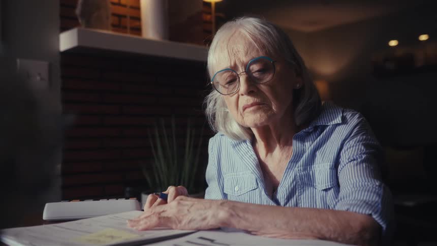 A concerned elderly woman with glasses sits at her table in a dimly lit room, carefully reviewing her bills and finances with a worried expression