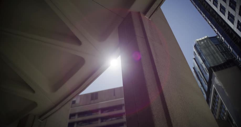 A low angle shot of the sun slowly emerging from behind a concrete pillar, creating a large lens flare against a modern city skyline