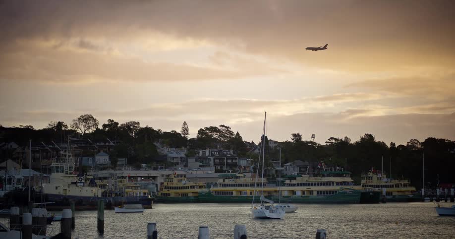 An airplane flies over a tranquil harbor with docked ferries and sailboats against a golden sunset sky