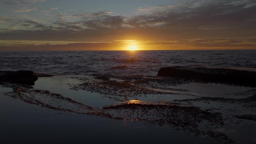 A serene sunrise over the ocean is reflected in the calm waters of a rocky tide pool
