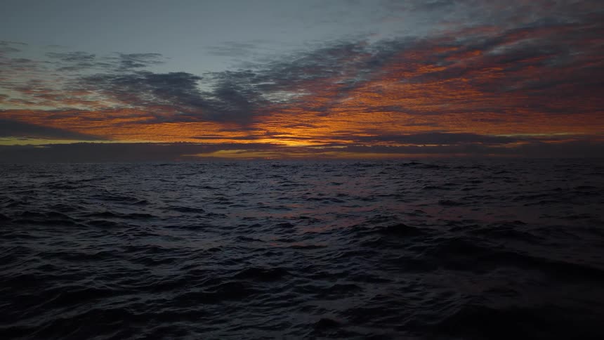 A dramatic orange and red sunset reflects on the gentle waves of the dark, open ocean