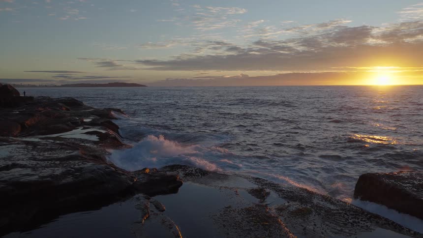 Slow-motion waves crash against the rocky shoreline, illuminated by the golden light of the rising sun over the ocean
