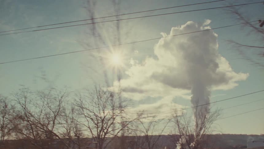 A nostalgic and hazy slow-motion drive-by view of a factory smokestack emitting steam into the bright sunlit sky