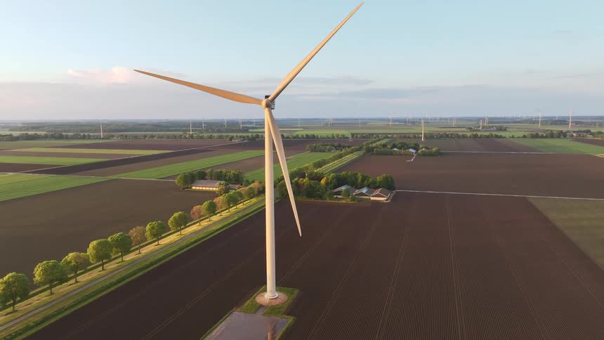 Static aerial view of a wind turbine slowly turning amidst vast agricultural fields at sunset