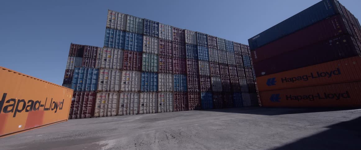 A low-angle slow pan reveals towering stacks of multicolored shipping containers at a large industrial port on a sunny day
