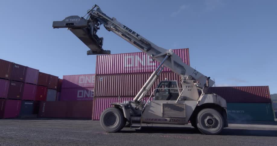 A large Terex reach stacker backs away from a stack of magenta shipping containers in an industrial port yard on a sunny day