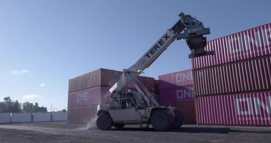 A large industrial reach stacker slowly moves into position to lift a shipping container from a stack in a port yard under a clear blue sky