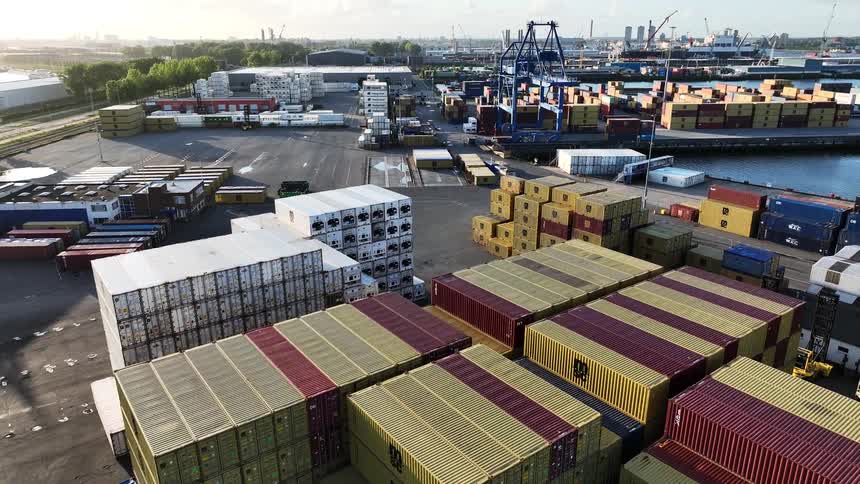 An ascending aerial shot over stacks of colorful shipping containers reveals a large industrial harbor with cranes and a city skyline in the distance