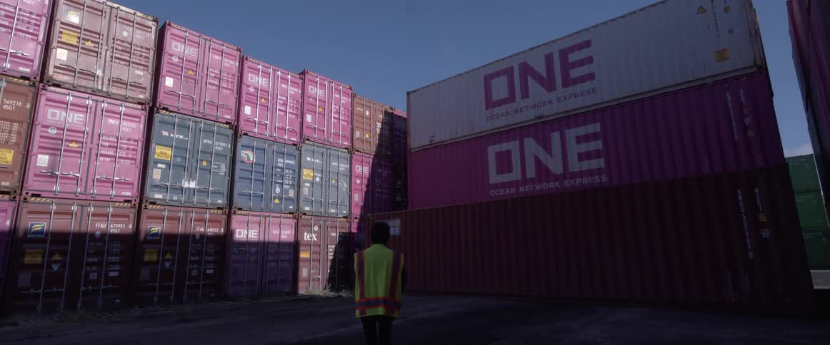 A worker in a high-visibility vest walks through a shipping yard with tall stacks of colorful cargo containers