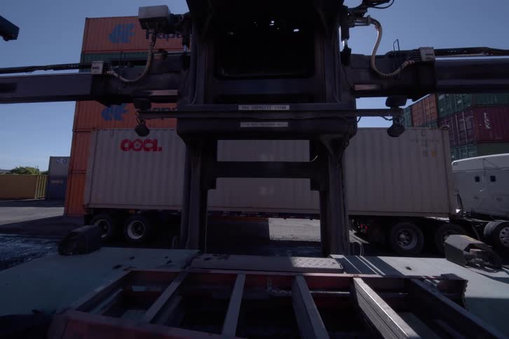 A time-lapse view from a large forklift shows shadows lengthening as it moves towards a container on a truck in a shipping yard