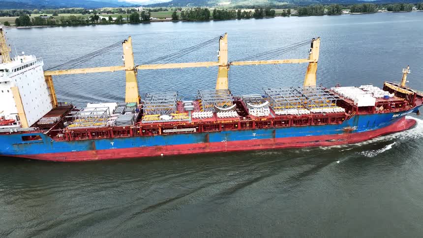 Aerial drone footage follows a large, colorful industrial cargo ship as it sails along a wide river with a green, rural shoreline in the distance
