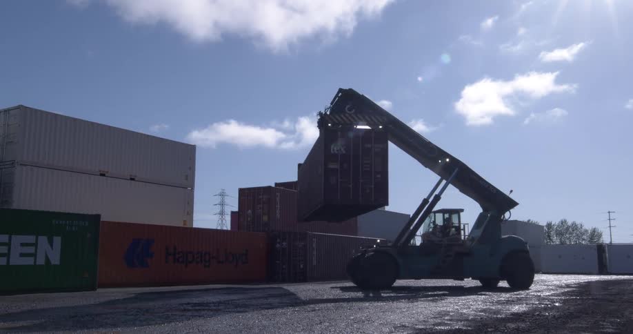 A reach stacker moves slowly across a shipping yard before lifting and stacking a large cargo container under a bright sun