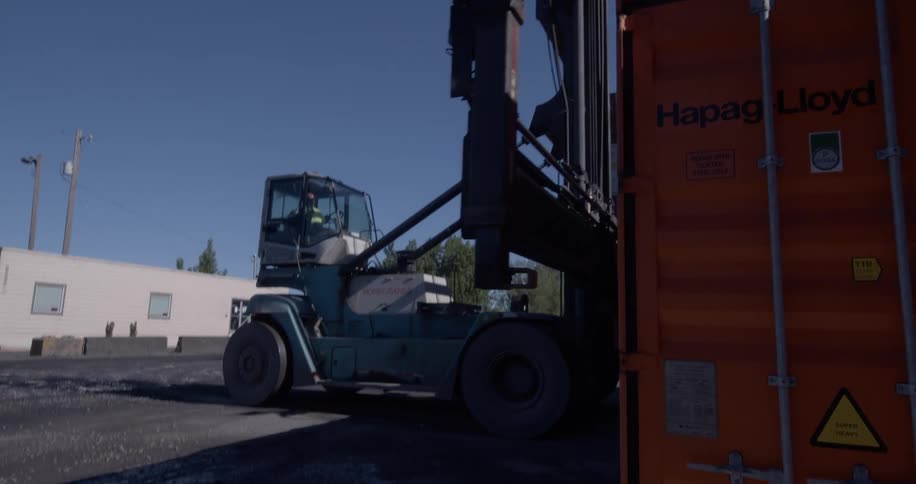 A large industrial forklift lowers its arm next to an orange shipping container in a shipping yard on a sunny day