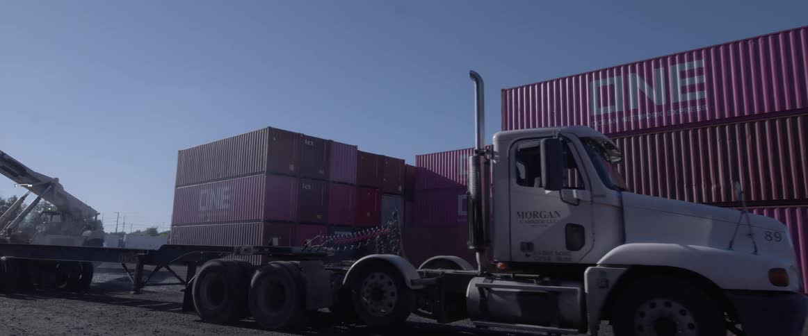 A large truck leaves a shipping yard after a crane unloads its cargo container