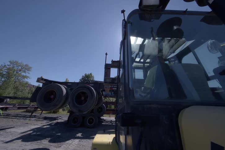 A close-up view from the side of a forklift operator skillfully lowering a semi-trailer chassis in a sunlit industrial yard