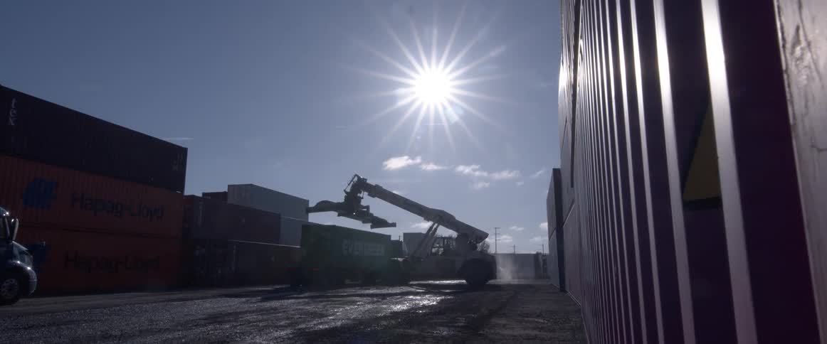 A reach stacker is silhouetted by the sun as it loads a shipping container onto a truck in an industrial yard