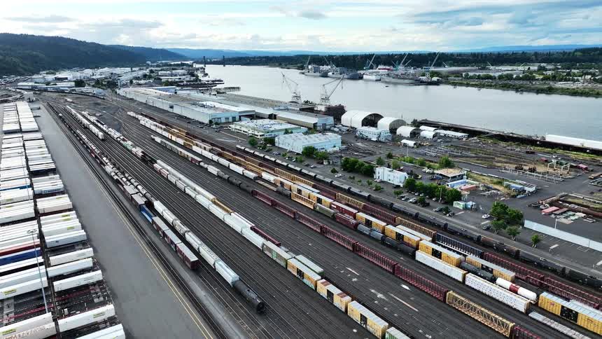 A static aerial view shows a long tanker train slowly moving through a vast industrial railyard and shipping port
