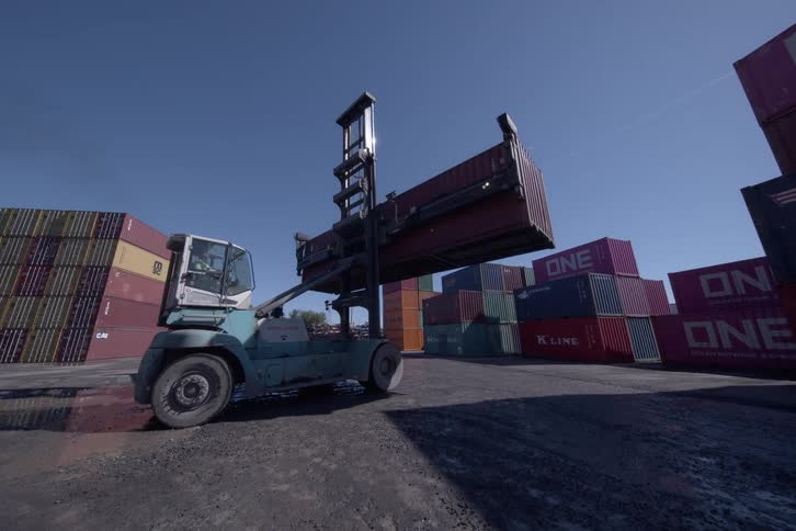 A top loader forklift lifts a shipping container off a semi-truck and reverses through a large industrial container yard on a sunny day