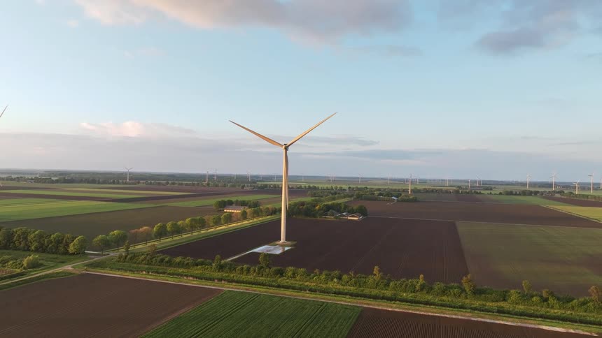Slow aerial pull-back shot of a large wind turbine in a vast rural landscape with agricultural fields and a wind farm at sunset