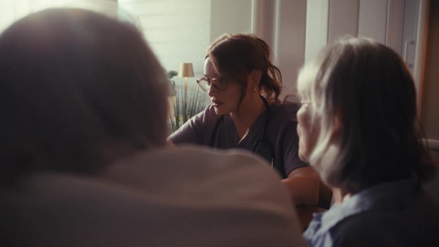 An over-the-shoulder view of a compassionate young nurse consulting with an elderly couple during an in-home visit