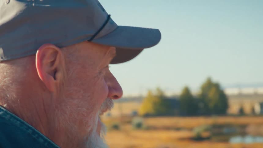 A thoughtful older man with a white beard and cap watches a freight train pass through the countryside