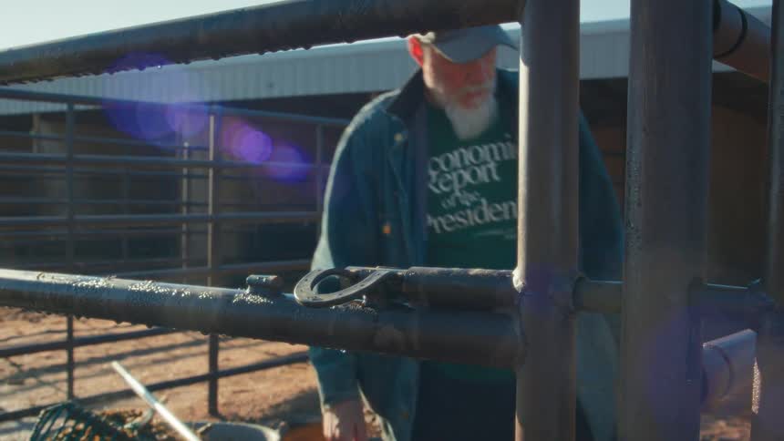 A bearded farmer finishes cleaning a horse stall and pauses at the gate to handle a horseshoe on a sunny ranch