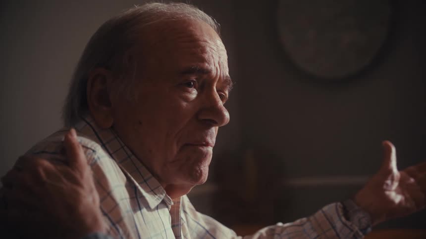A close-up, slow-motion shot captures an elderly man in an emotional, heartfelt conversation as a supportive hand rests on his shoulder