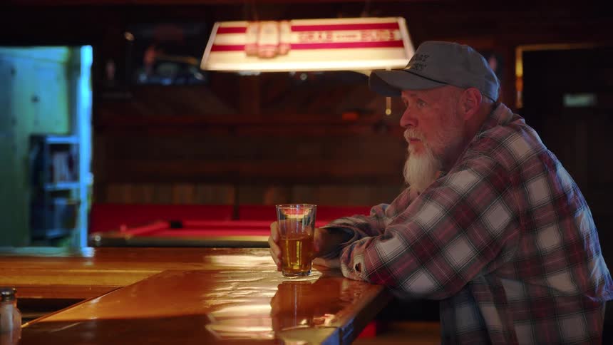A contemplative senior man with a long white beard sits alone and drinks a beer at a rustic wooden bar
