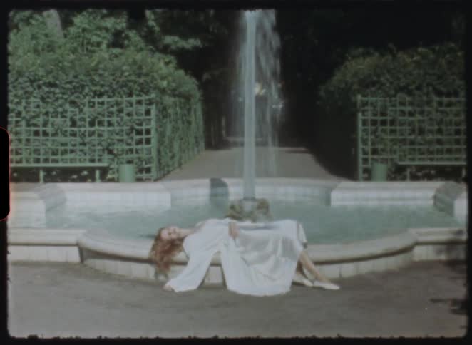 A dreamy vintage shot slowly zooms in on a serene woman with red hair in a white gown, resting by a beautiful garden fountain