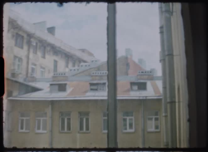 Vintage 8mm footage zooms in from a window onto the weathered facade and rooftops of an old European apartment building on an overcast day