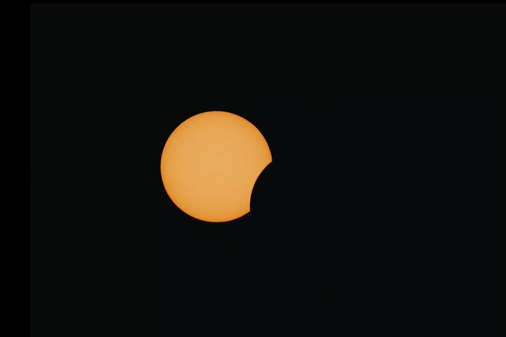 A close-up, real-time view of a partial solar eclipse, showing the moon slowly covering the bright orange sun against a black sky