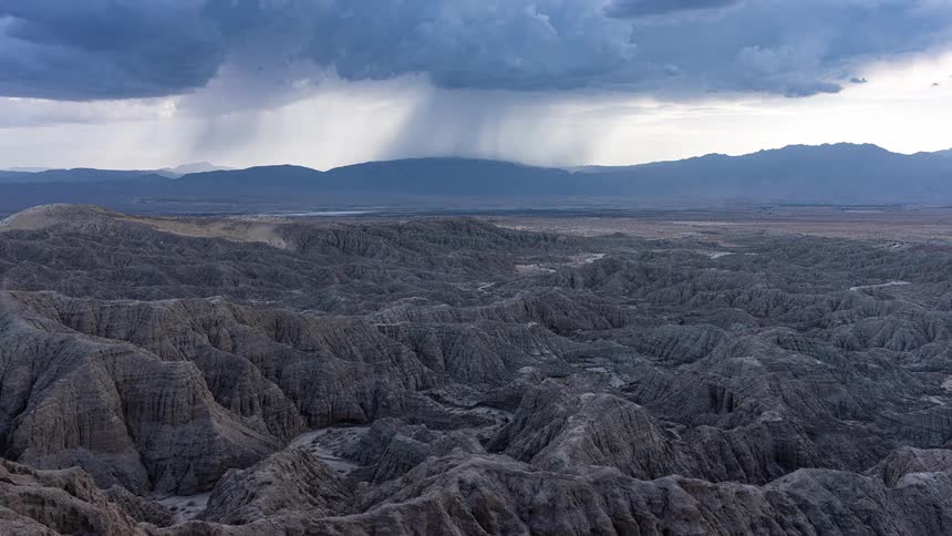 An ominous storm unleashes a downpour over mountains, viewed across a desolate and eroded badlands landscape in this cinemagraph