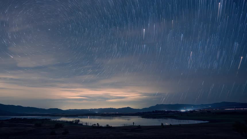 A stunning timelapse shows star trails rotating in the night sky over a calm lake and distant mountains
