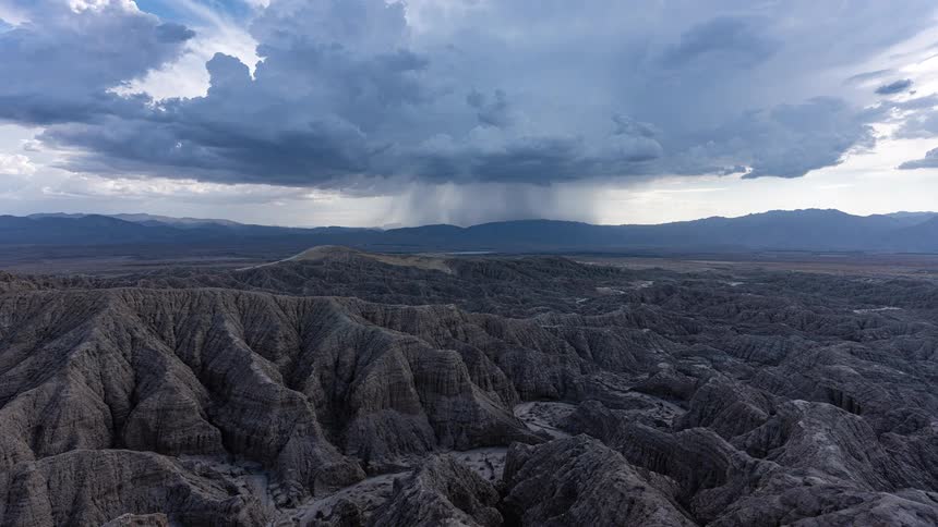 A dramatic time-lapse captures a dark, swirling storm cloud releasing a downpour over a vast and rugged badlands landscape