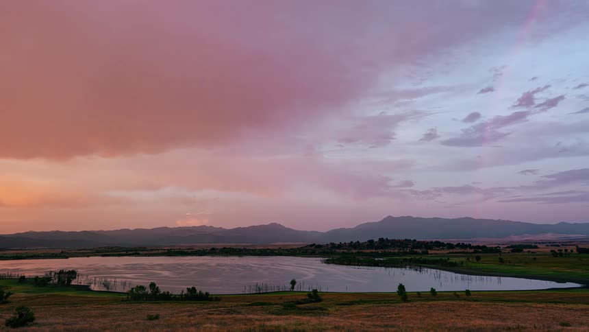 A stunning timelapse of a vibrant pink and orange sunset with a rainbow appearing over a mountain lake