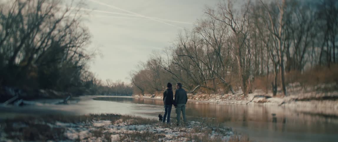 From behind, a couple and their dog enjoy the peaceful view of a river surrounded by a snowy, wooded landscape in winter