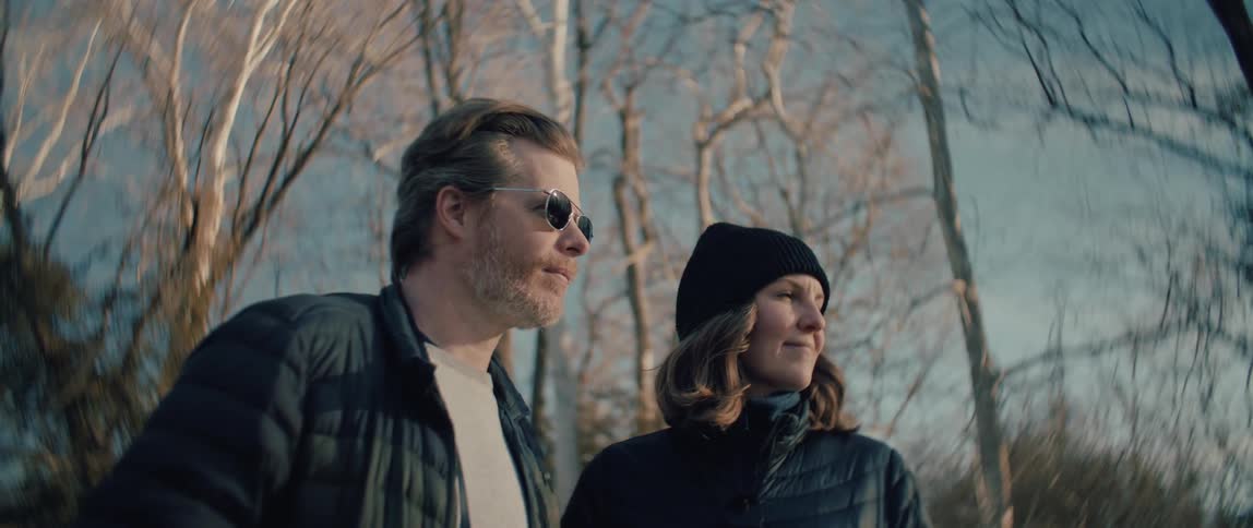 A couple wearing winter jackets stands side-by-side in the woods, peacefully admiring the view together