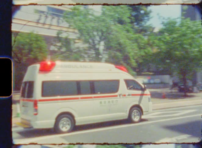 A Tokyo Fire Department ambulance with flashing lights drives down a city street in Japan, captured on vintage 8mm film