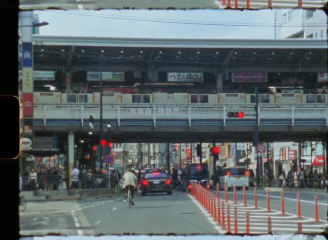 A vintage Super 8 film captures the daily hustle of a Japanese city street with cyclists, cars, and an elevated train passing by