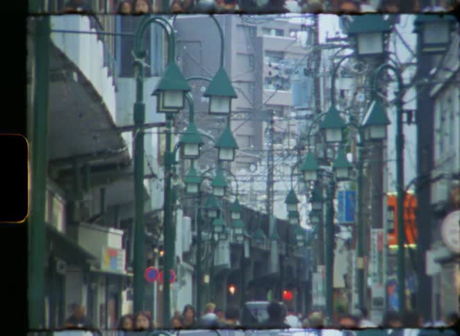 Vintage 8mm footage captures a bustling street scene in Japan, with pedestrians walking under rows of green lanterns and elevated train tracks