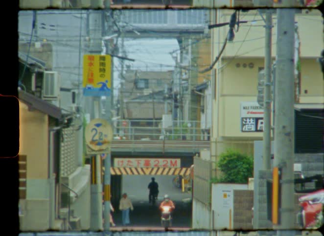 Vintage 8mm film captures pedestrians, a cyclist, and a scooter passing through a low underpass on a narrow urban street in Japan