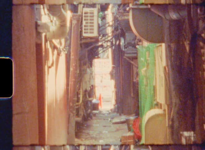 An 8mm film aesthetic captures a static, experimental shot down a narrow and cluttered urban alleyway