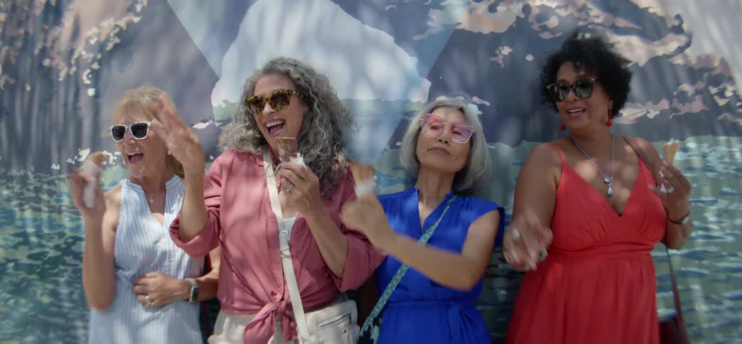 A diverse group of fun-loving senior women laugh and dance while eating ice cream cones on a sunny day