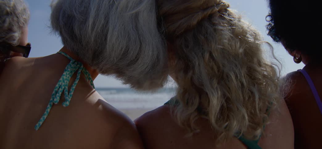 A close-up from behind of three diverse female friends sitting together on the beach, looking out at the ocean on a sunny day