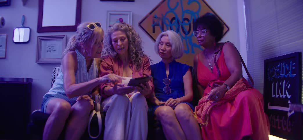A diverse group of four stylish senior women sit together on a couch, talking and sharing something on a tablet in a room with ambient purple light