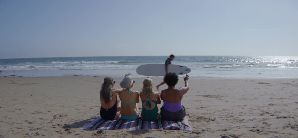 Four senior women sitting on a blanket at the beach wave to a passing surfer on a sunny day