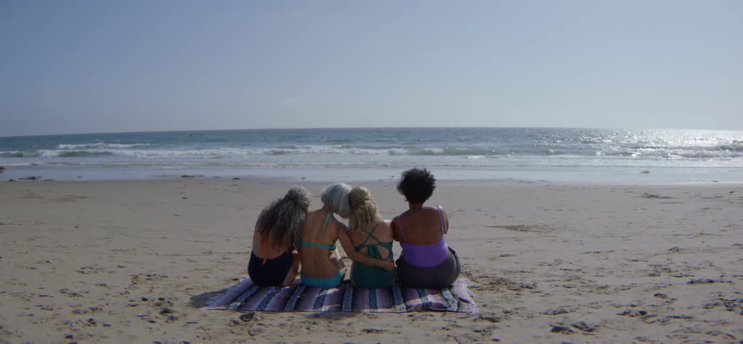 A group of older female friends sits together on a beach blanket, embracing and watching the ocean on a sunny day