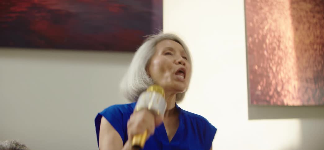 An enthusiastic elderly woman with grey hair belts out a song on a karaoke microphone, enjoying a lively party with friends in her home