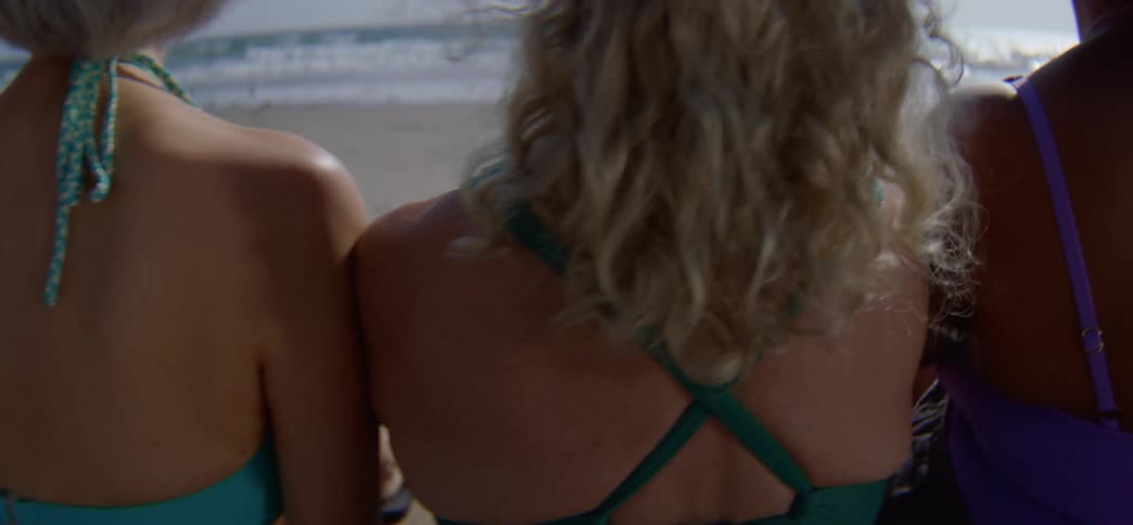 Close up from behind on a group of diverse women in swimsuits as they sit shoulder-to-shoulder on the beach watching the ocean